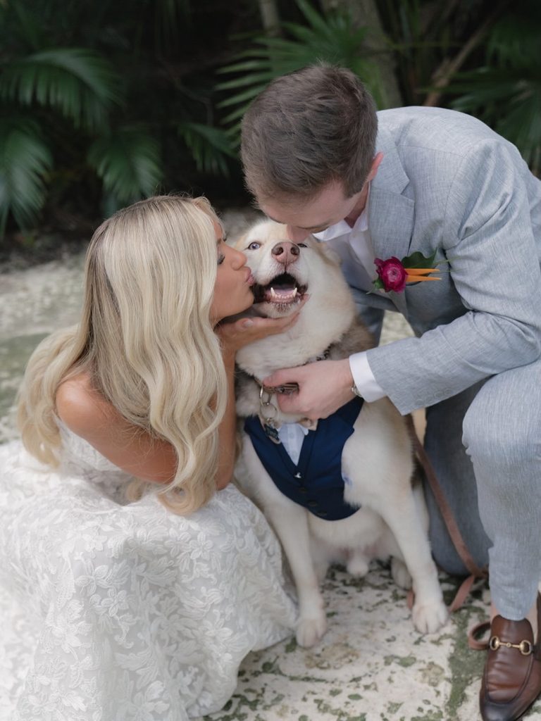 Bride and groom kissing their happy husky wearing navy vest with tropical foliage behind them