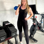 Madeline holding blue kettlebell in home gym with exercise bike and weights