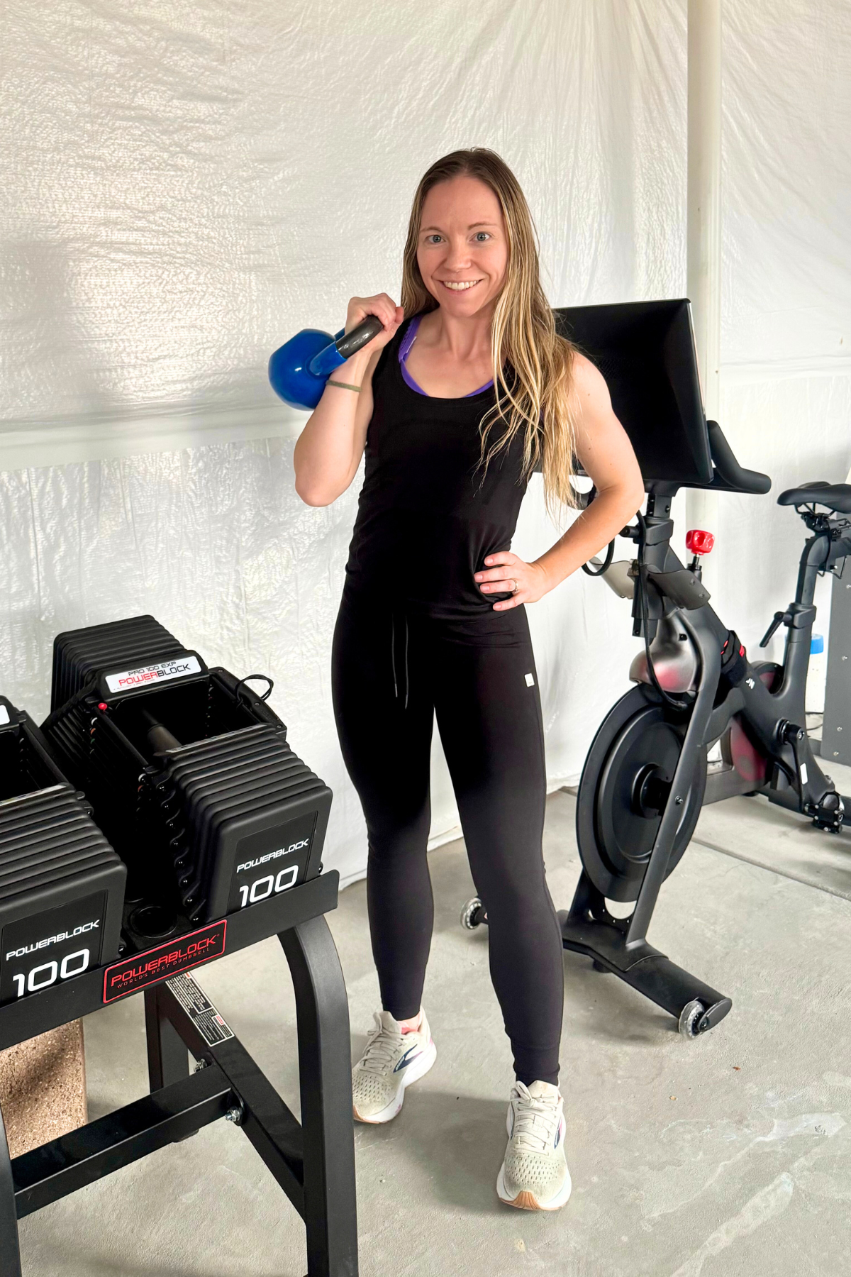 Madeline holding blue kettlebell in home gym with exercise bike and weights