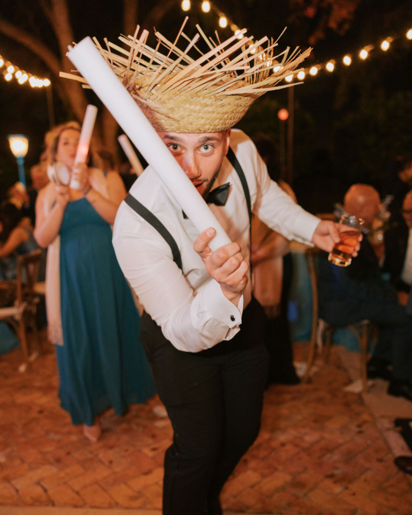 Guest wearing straw hat celebrates with foam sticks during outdoor reception under string lights