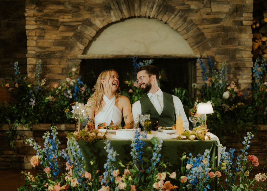 Mallory and Andrew laugh together at sweetheart table decorated with blue and peach florals in front of stone fireplace