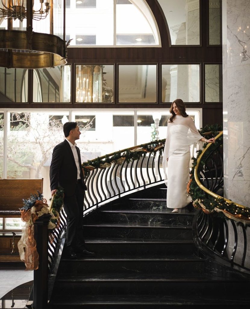 Bride descends hotel's grand black marble staircase with ornate railings while groom waits below