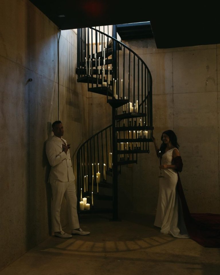 Bride and groom in white attire standing on opposite sides of illuminated spiral staircase with candles