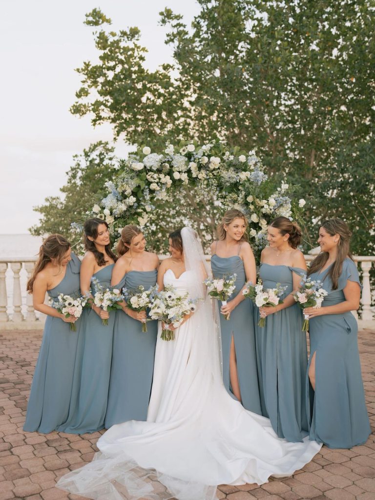 Bride in white gown with bridesmaids in dusty blue dresses holding white bouquets beneath floral arch at waterfront ceremony