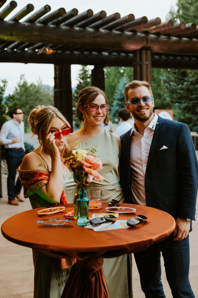 Three wedding guests wearing retro sunglasses pose at cocktail table under wooden pergola at outdoor mountain reception