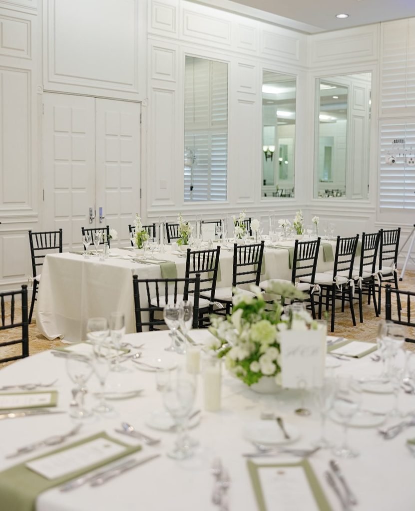 Elegant white reception room with round tables, black chiavari chairs, and green accent napkins
