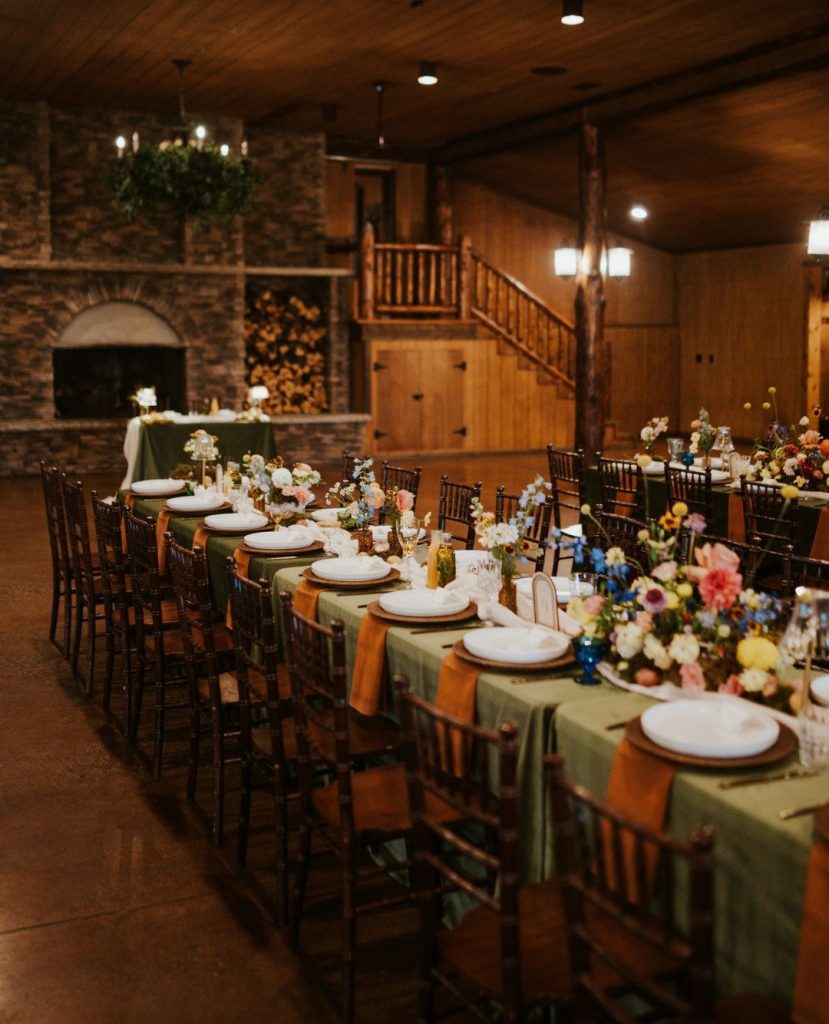 Long reception tables with sage green runners, colorful floral centerpieces, and dark wooden chairs inside rustic stone lodge