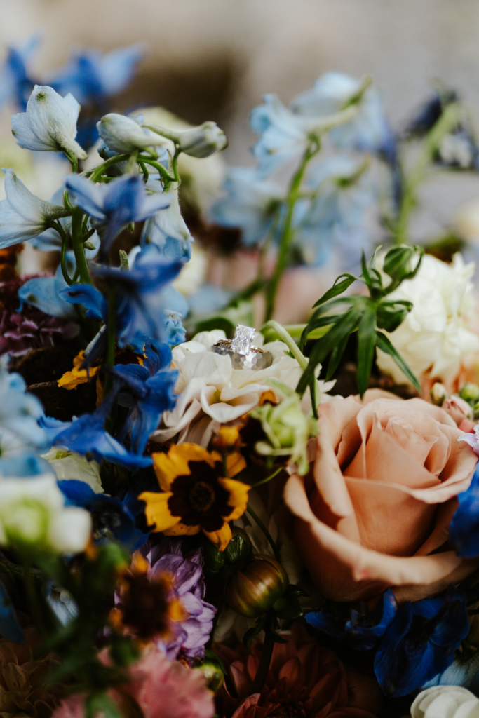 Close-up of bridal bouquet featuring blue delphinium, peach roses, sunflowers, and diamond engagement ring nestled in blooms