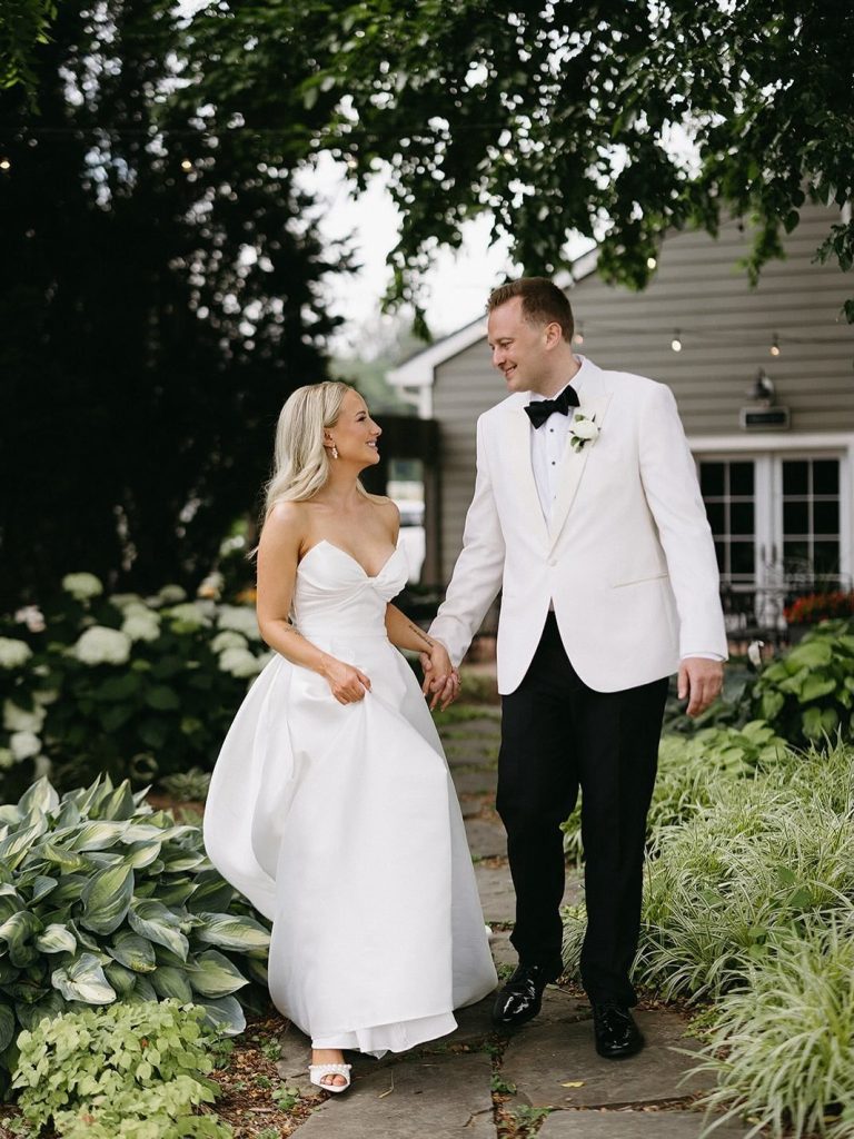 Bride and groom holding hands while walking through garden path lined with hydrangeas and hostas