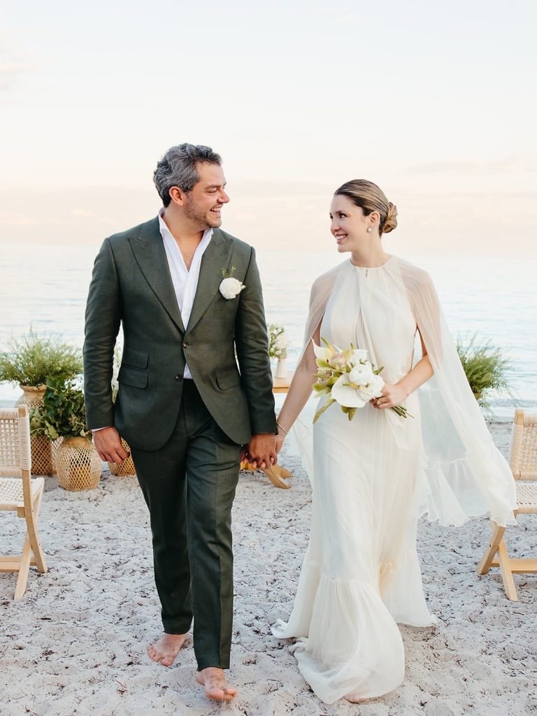 Newlyweds hold hands walking barefoot on beach at sunset ceremony