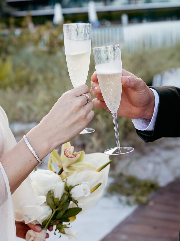 Bride and groom toasting with champagne flutes, bridal bouquet in hand at waterfront ceremony