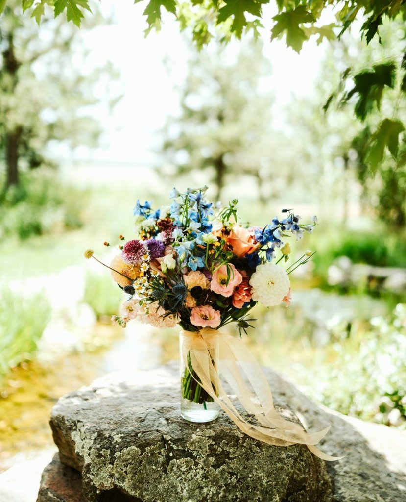 Wildflower bridal bouquet with blue delphinium, pink poppies, and burgundy blooms resting on stone wall outdoors