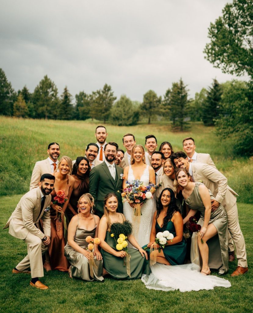 Wedding party of twenty people in sage, terracotta, and cream attire posed together on lawn with mountain landscape behind