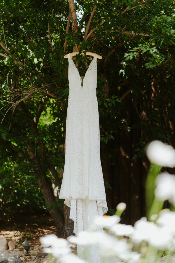 White lace wedding dress with plunging V-neckline hanging from tree branch surrounded by foliage and white flowers