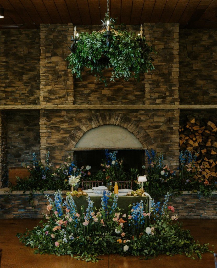 Sweetheart table with abundant blue delphinium and peach rose arrangements against rustic stone fireplace with greenery chandelier above
