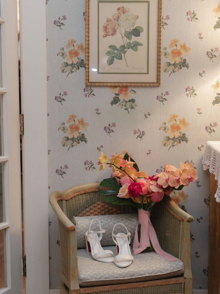 Bridal bouquet and white heeled sandals displayed on vintage wicker chair against floral wallpaper