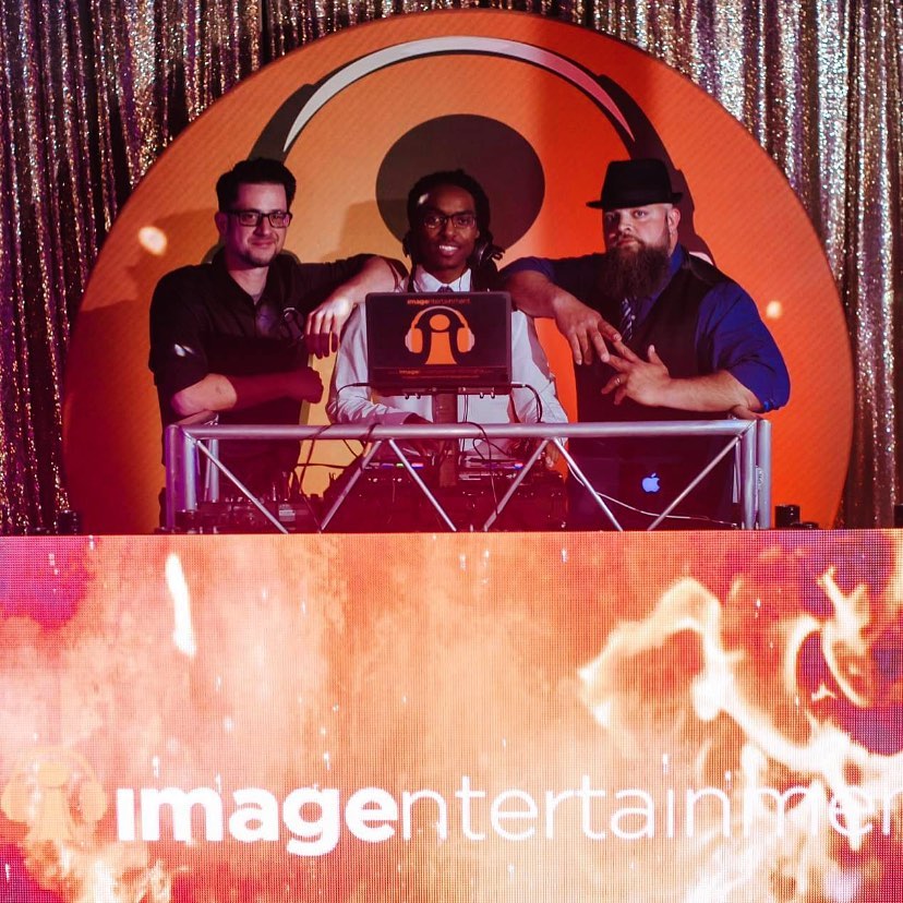 Three DJs posing behind booth with glittery backdrop and orange circular design at wedding