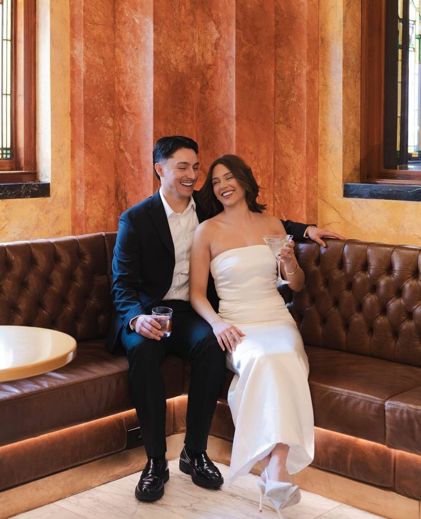 Newlyweds laughing together on leather sofa against ornate marble columns
