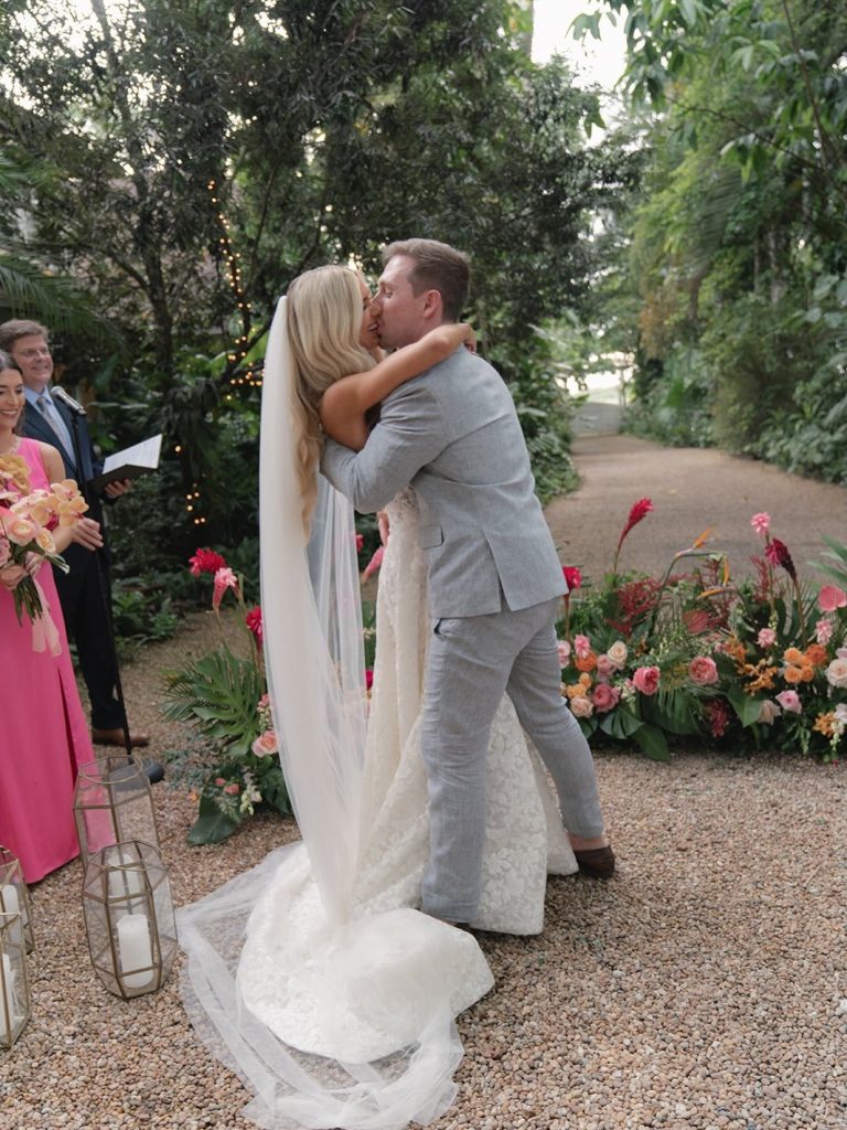 Newlyweds share their first kiss at an outdoor garden ceremony with colorful floral arrangements and string lights