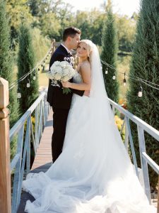 Newlyweds pose on bridge surrounded by lush greenery and romantic string lighting