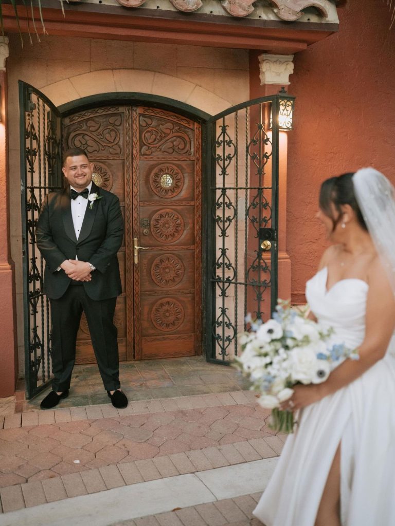 Groom smiles at bride during first look moment near carved wooden doors with wrought iron gates