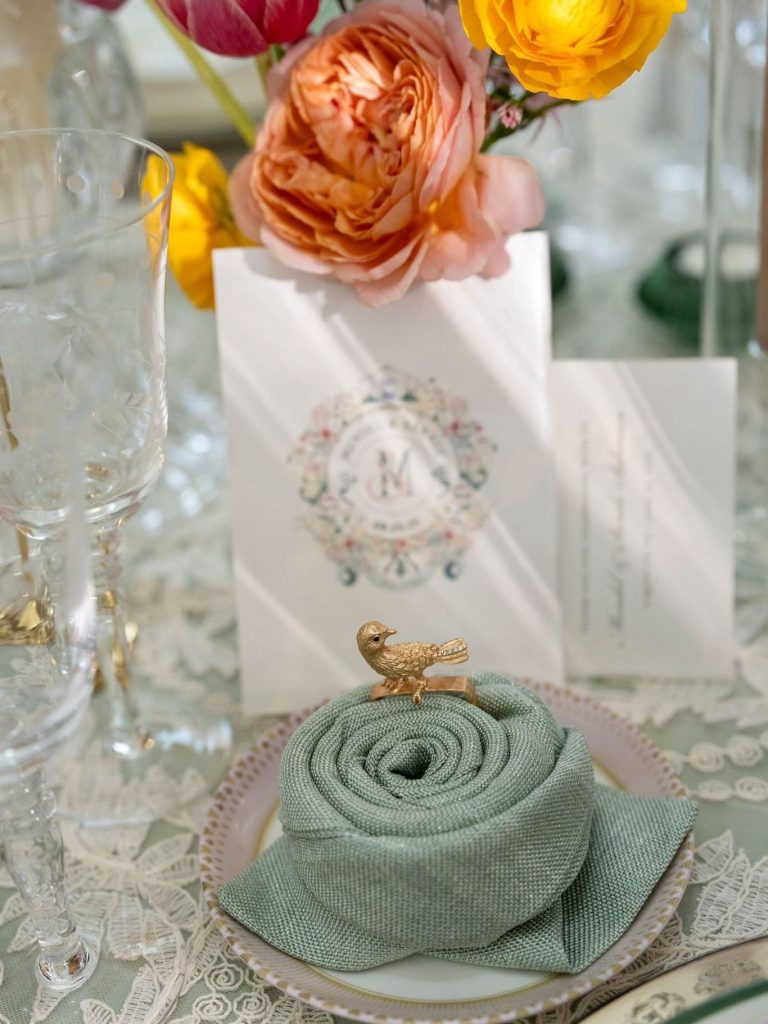 Elegant place setting features sage green rolled napkin with gold bird figurine, monogrammed menu, and colorful blooms