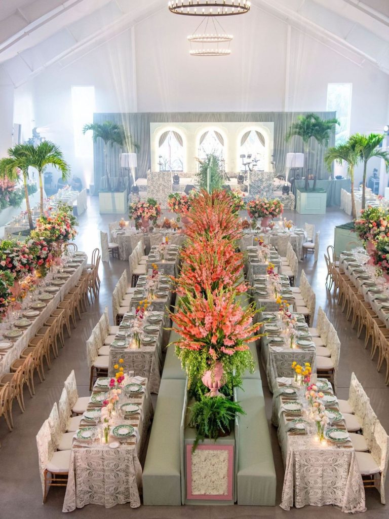 Grand reception hall with long tables, tropical centerpieces, and dramatic pink floral arrangements under vaulted ceiling