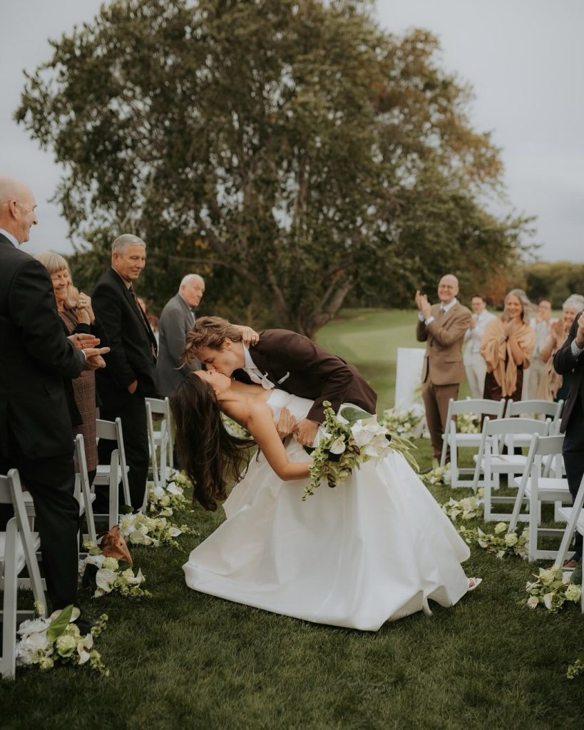 Bride and groom sharing romantic first kiss at outdoor ceremony with guests applauding beneath autumn trees