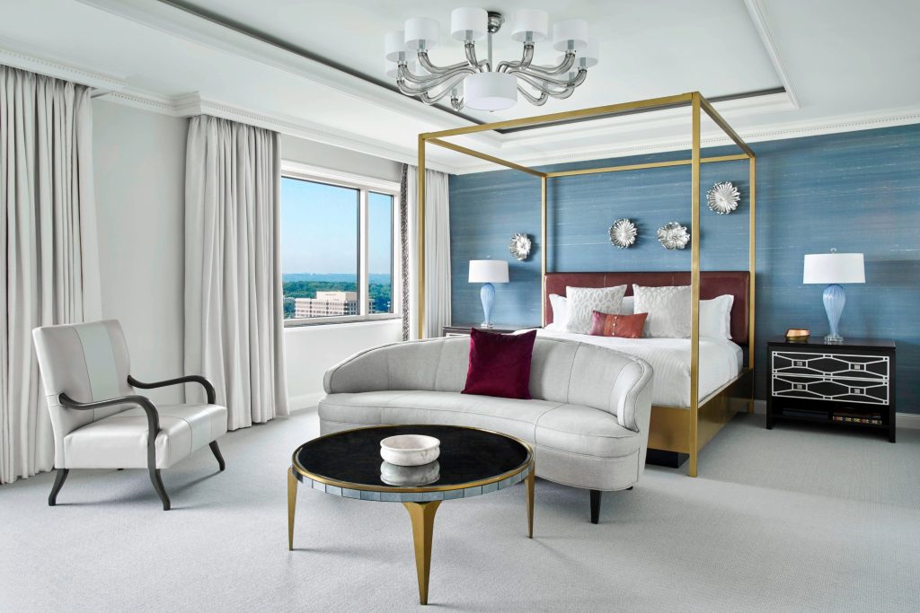 Luxurious hotel bedroom with gold canopy bed, curved white sofa, blue accent wall, and city views