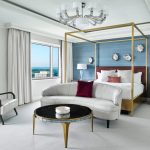 Luxurious hotel bedroom with gold canopy bed, curved white sofa, blue accent wall, and city views
