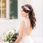 Bride in backless beaded wedding gown with half-up curled hairstyle and floral hair accessory holding greenery bouquet