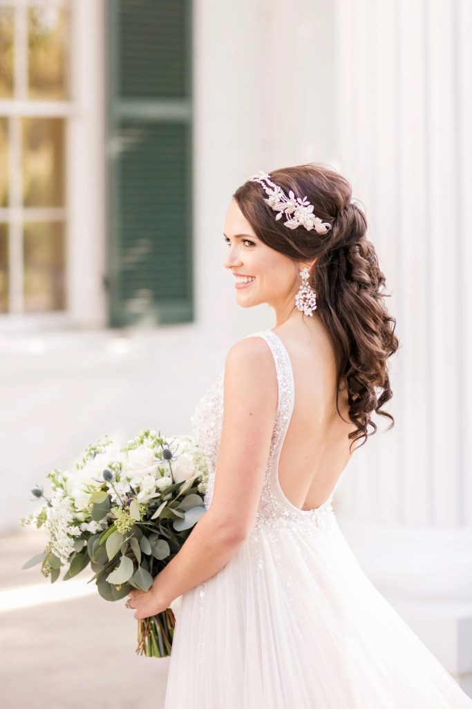 Bride in backless beaded wedding gown with half-up curled hairstyle and floral hair accessory holding greenery bouquet
