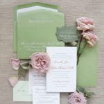 Sage green and blush pink wedding invitation suite with calligraphy addressing, arranged with pink roses and ribbon on neutral linen background