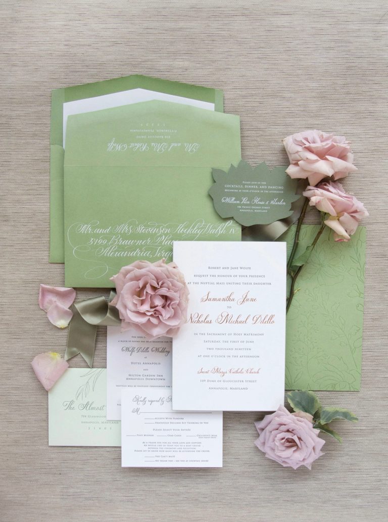 Sage green and blush pink wedding invitation suite with calligraphy addressing, arranged with pink roses and ribbon on neutral linen background