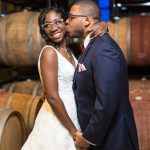 Groom kissing bride on the cheek in a barrel room, showcasing wedding hair and makeup styling