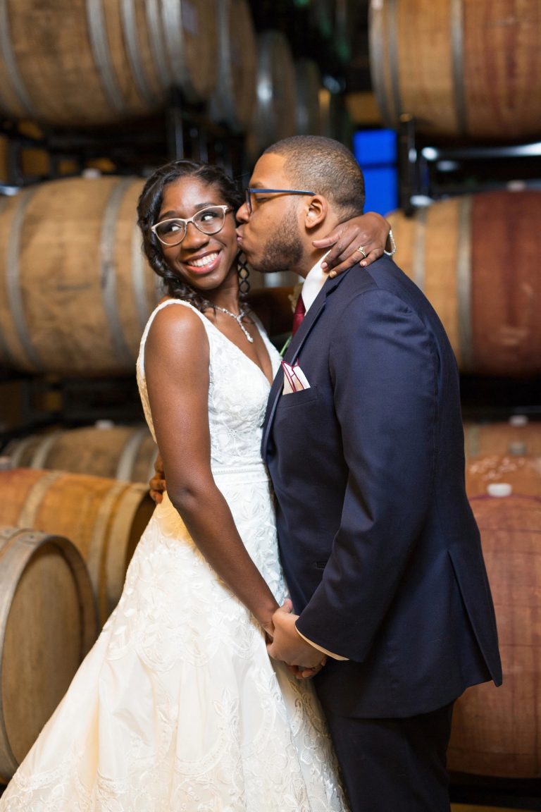 Groom kissing bride on the cheek in a barrel room, showcasing wedding hair and makeup styling