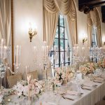 Luxurious wedding reception table with crystal candelabras, blush floral arrangements, and gold-trimmed place settings in ballroom with swag curtains