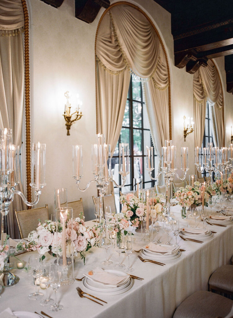 Luxurious wedding reception table with crystal candelabras, blush floral arrangements, and gold-trimmed place settings in ballroom with swag curtains