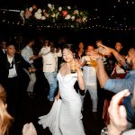 Bride dancing joyfully with wedding guests on illuminated dance floor under floral arch and string lights