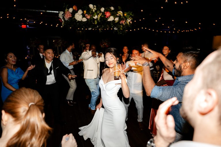 Bride dancing joyfully with wedding guests on illuminated dance floor under floral arch and string lights