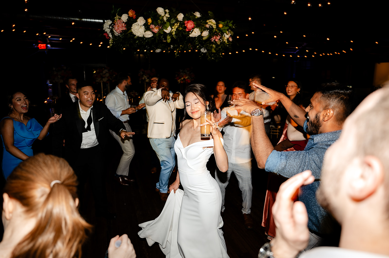 Bride dancing joyfully with wedding guests on illuminated dance floor under floral arch and string lights
