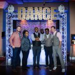 AMP'D Entertainment team poses beneath illuminated "DANCE" marquee sign with professional lighting setup at brick venue