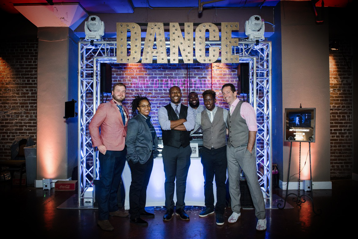 AMP'D Entertainment team poses beneath illuminated "DANCE" marquee sign with professional lighting setup at brick venue
