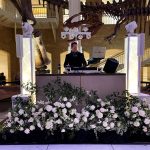 DJ booth setup at natural history museum with white floral arrangements and dinosaur skeleton displays