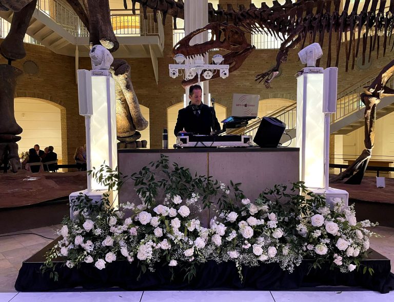 DJ booth setup at natural history museum with white floral arrangements and dinosaur skeleton displays