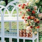 Outdoor wedding bar with white shelving displaying vibrant coral and pink floral arrangement, copper mugs, and glassware in garden setting
