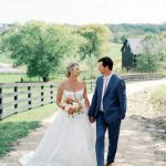 Bride in white ball gown holding coral and white bouquet walks hand-in-hand with groom in navy suit on rustic farm path with black barn and rolling hills in background.