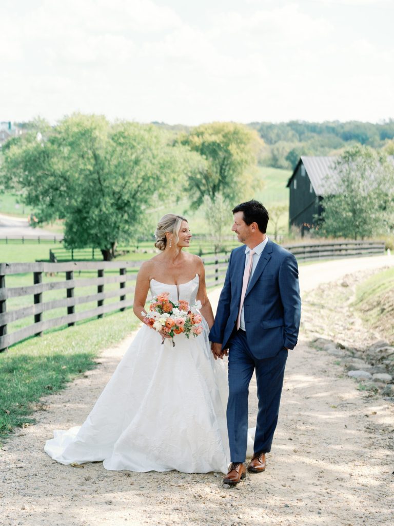 Bride in white ball gown holding coral and white bouquet walks hand-in-hand with groom in navy suit on rustic farm path with black barn and rolling hills in background.