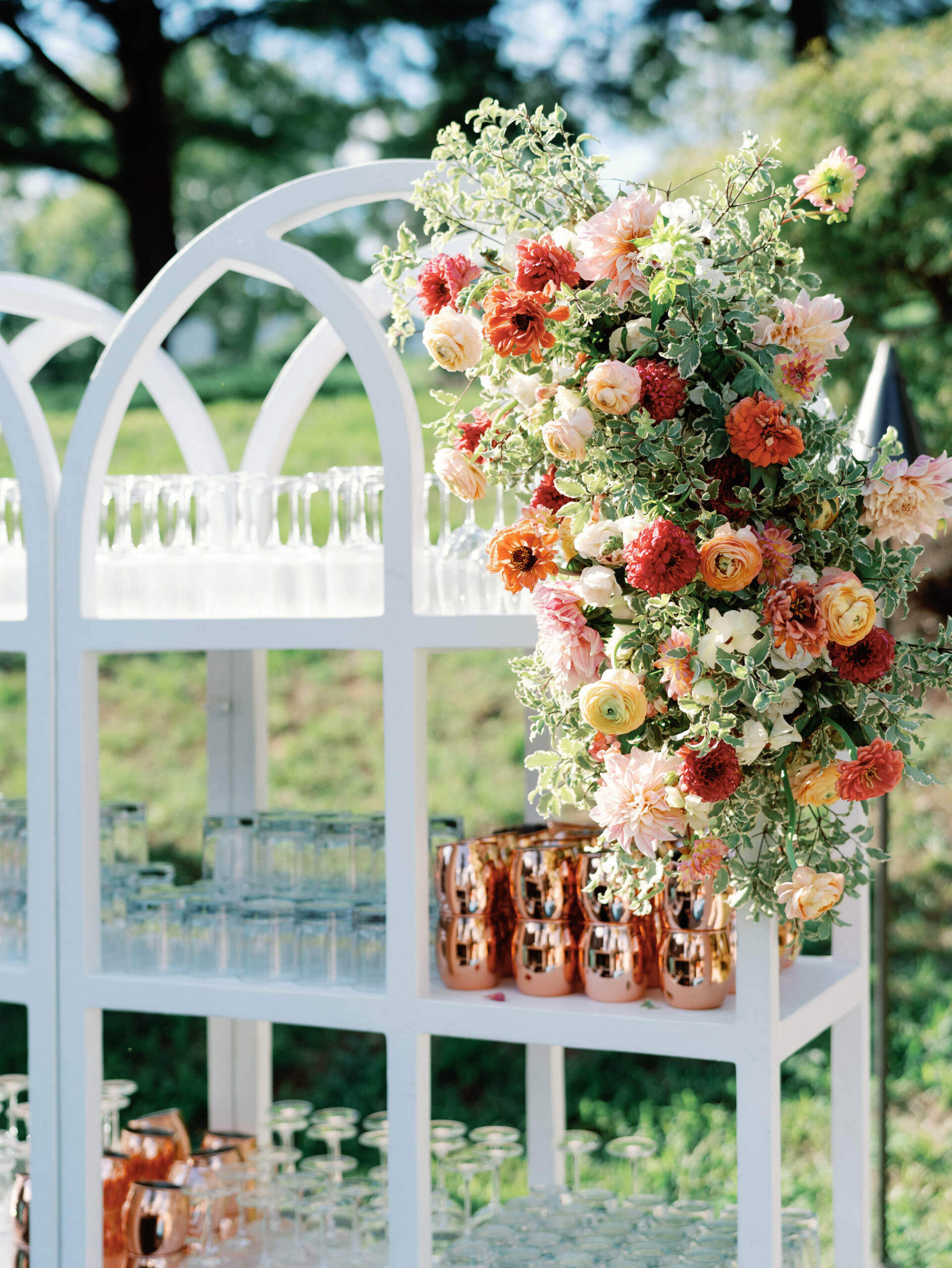 Outdoor wedding bar with white shelving displaying vibrant coral and pink floral arrangement, copper mugs, and glassware in garden setting