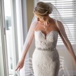 Bride in strapless lace wedding gown with crystal belt and cathedral veil, styled hair in elegant updo with jeweled headpiece
